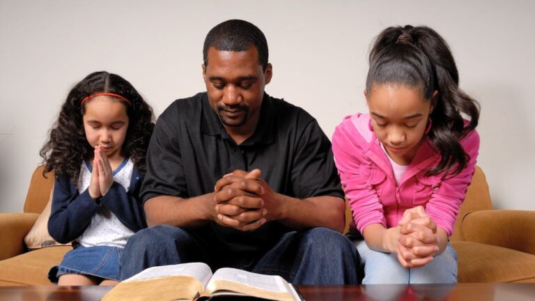 A father and his two daughters sitting on a couch, sharing a family prayer with their eyes closed and hands clasped together over a Bible.