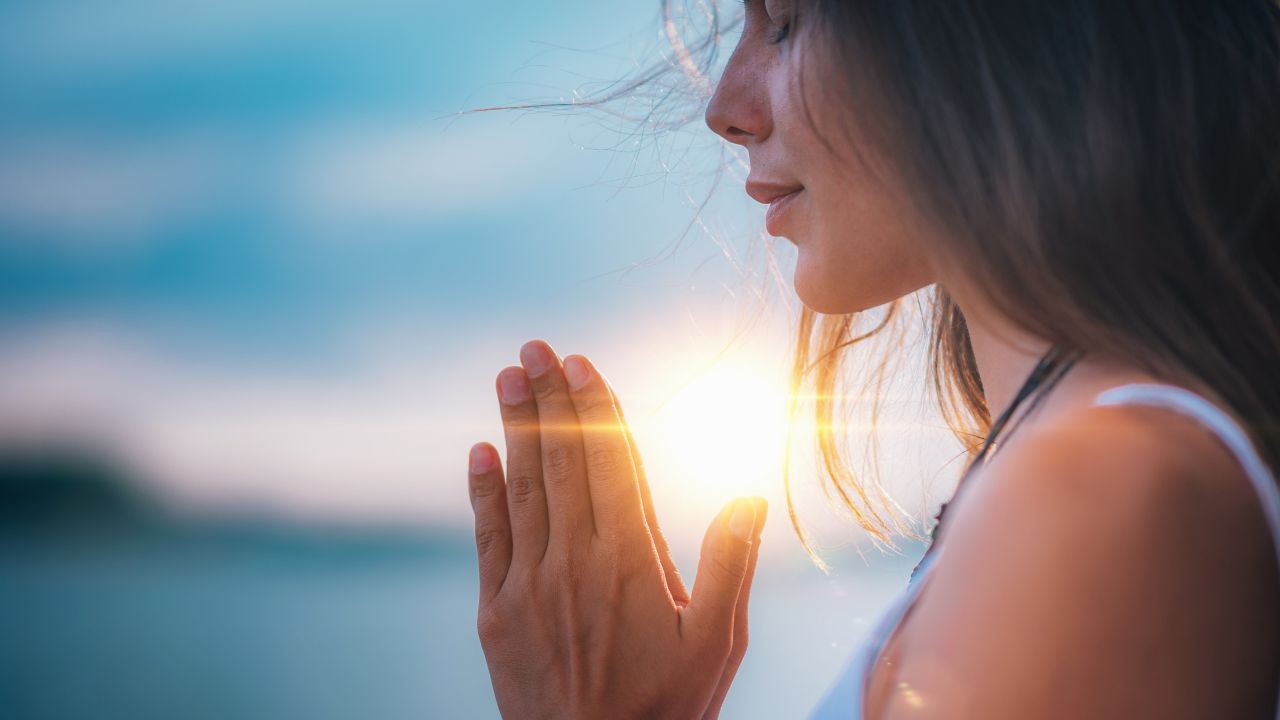 A woman meditating outdoors at sunrise, with sunlight shining through her hands as she gently holds them together in a prayer position.