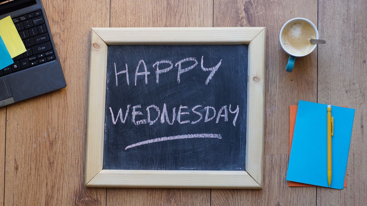 A chalkboard with the phrase "happy Wednesday blessings" written in white chalk, surrounded by a laptop, a cup of coffee, and colorful notebooks on a wooden table.