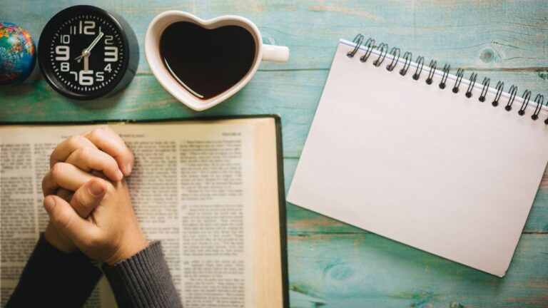A person's clasped hands resting beside an open Bible, a heart-shaped cup of coffee, a notebook, and a round clock on a wooden table.
