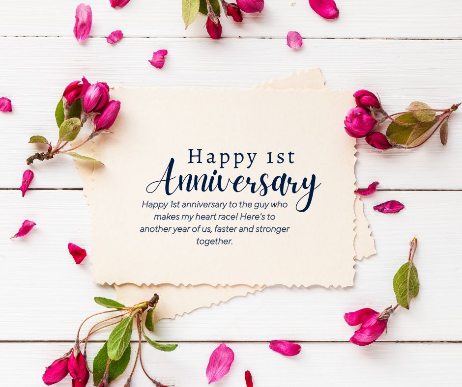 A romantic 1st anniversary card surrounded by pink flower petals on a white wooden background. The card reads "Happy 1st Anniversary Wishes" with a heartfelt message underneath.