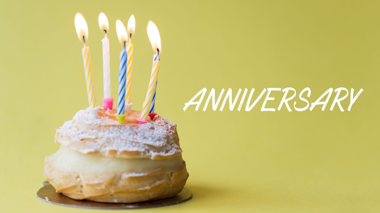 A cream-filled pastry topped with powdered sugar and four lit candles, with the words "happy anniversary" in bold, set against a soft yellow background.