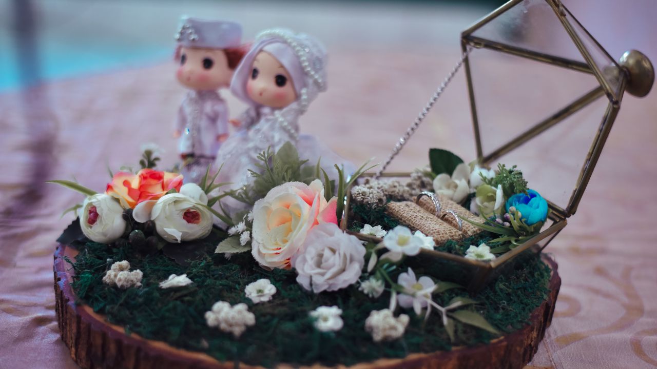 A decorative display featuring two small figurines dressed in wedding attire on a wooden slab with white and pink flowers, greenery, and a geometric metal ornament, perfect for wedding anniversary wishes for your wife.