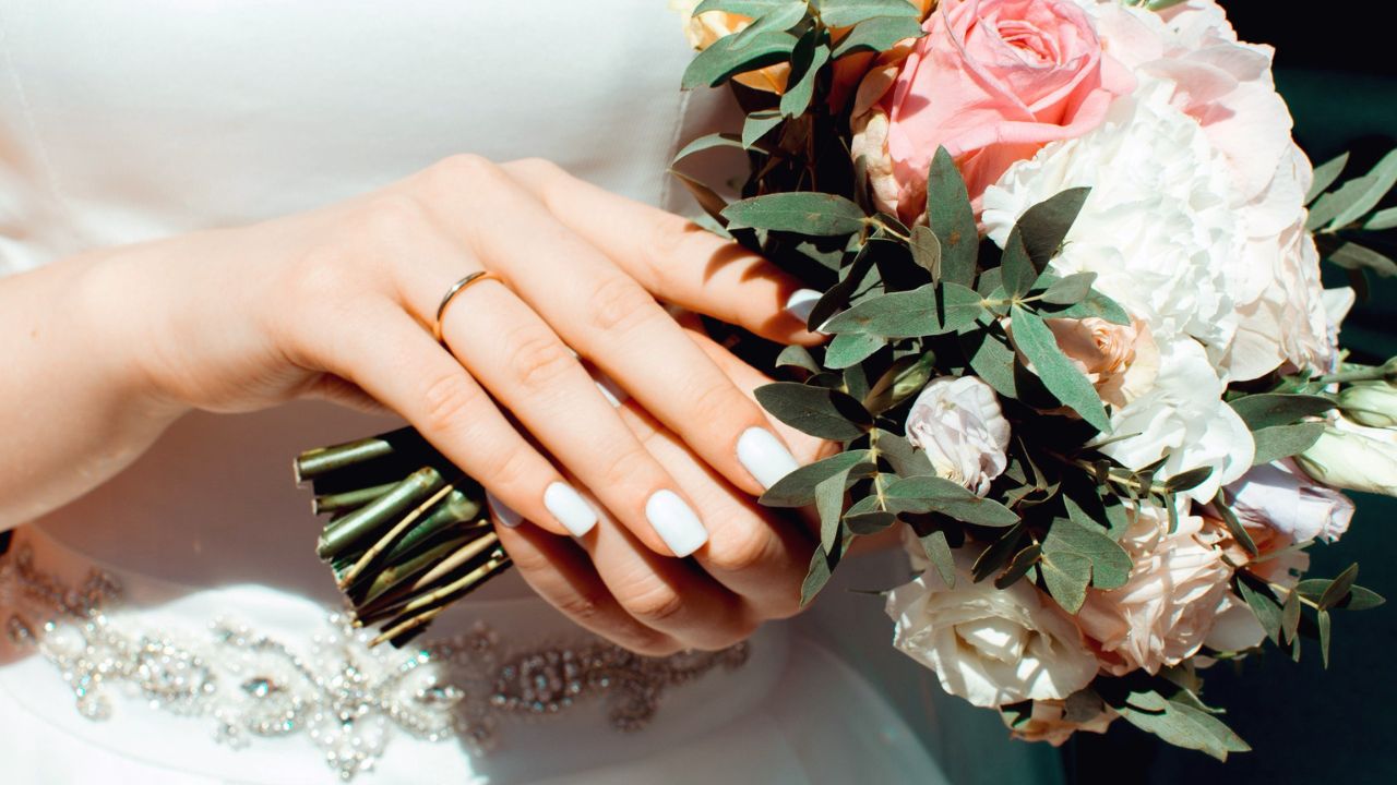 A close-up of a bride's hand holding a bouquet of pink and white flowers, featuring a prominent engagement ring and manicured nails, with a glimpse of her white dress and beaded accessory.