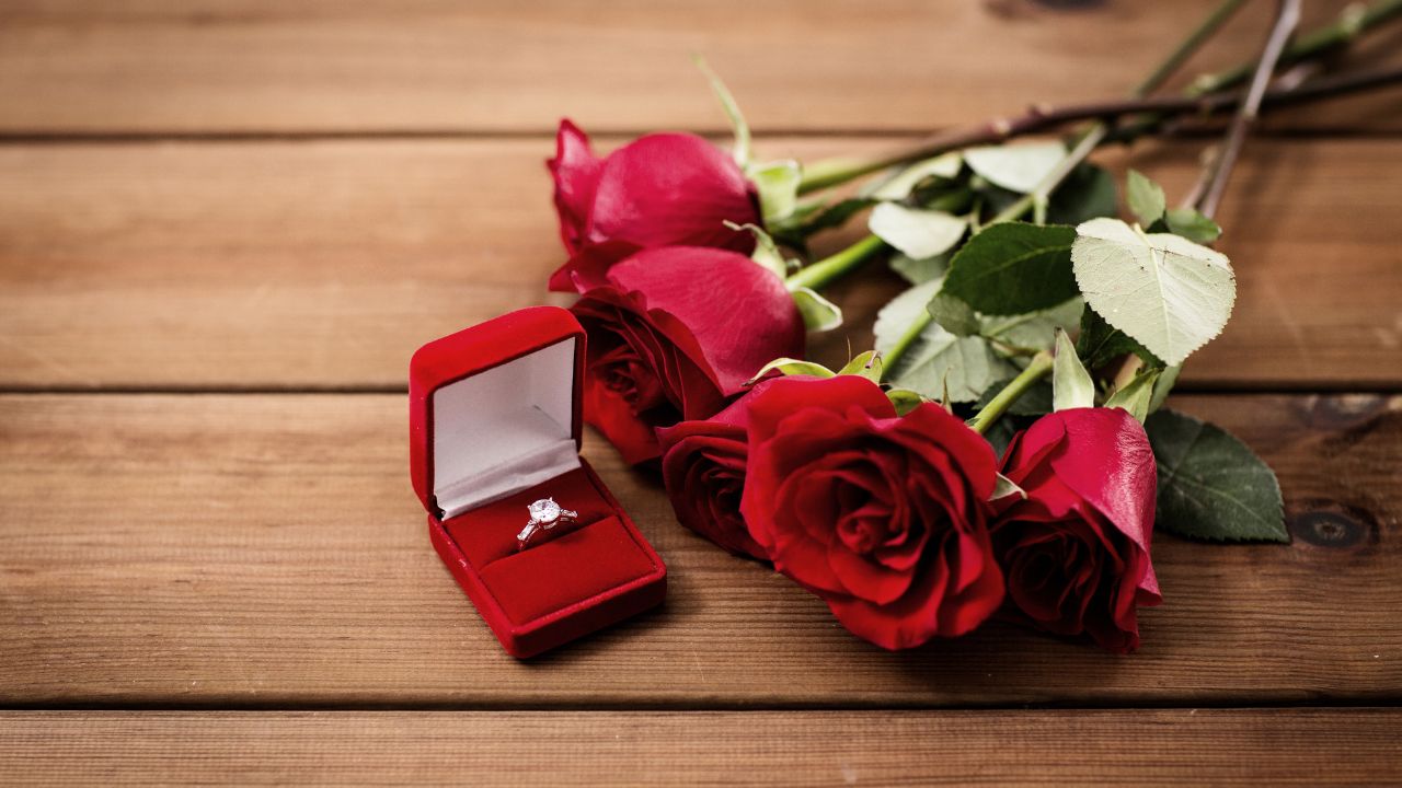 Three red roses lie on a wooden surface next to an open red box displaying an engagement ring, symbolizing love blooms.