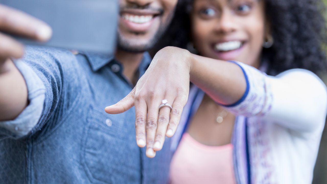 A joyful couple taking a selfie to showcase the woman’s happy engagement ring. The focus is on the ring as they smile broadly, capturing a moment of excitement and love.