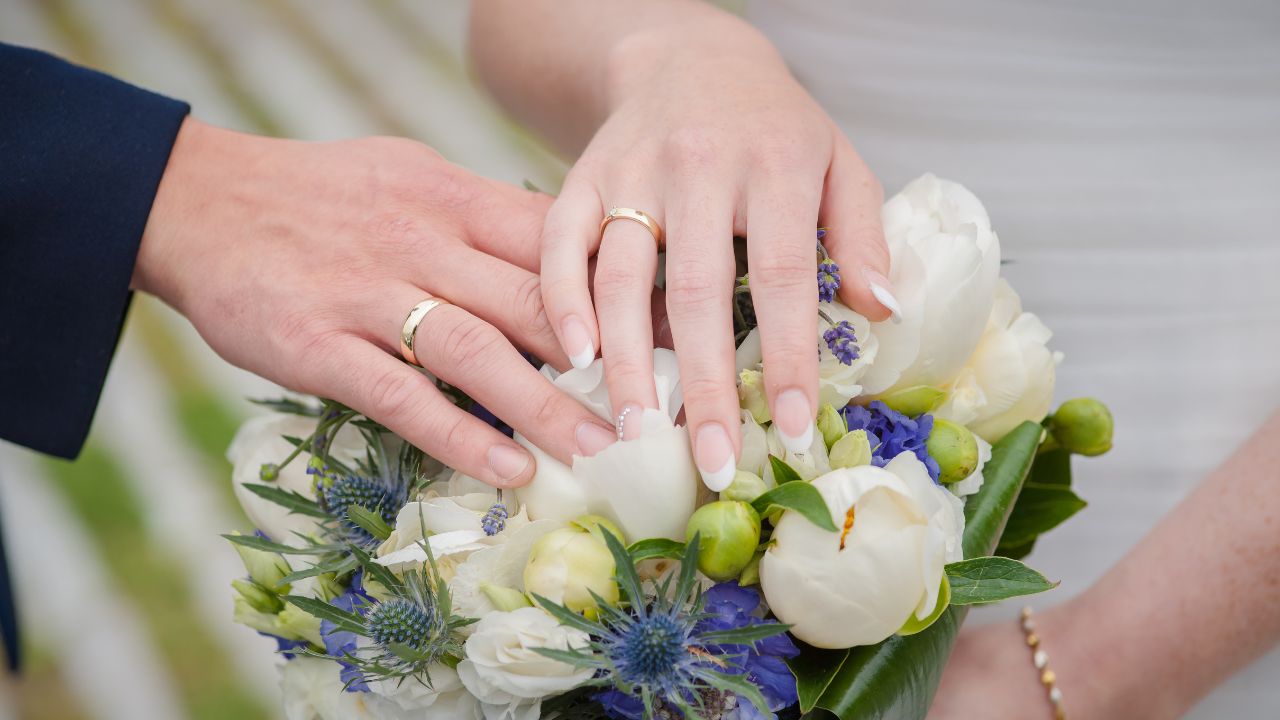 Close-up of a newly engaged couple's hands with engagement rings, placed over a bouquet of white and blue flowers.