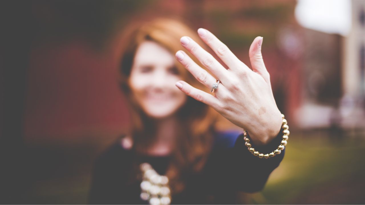A woman in soft focus smiles in the background, showing off her engagement ring by holding her hand up to the camera, with a clear view of the sparkling ring and her bracelet as love blooms around her