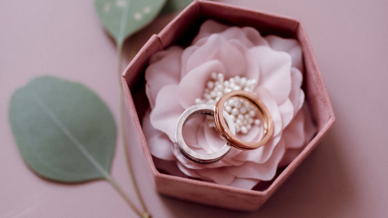 Two engagement rings on a pink flower inside a small pink hexagonal box with a green leaf beside it, set against a soft pink background.
