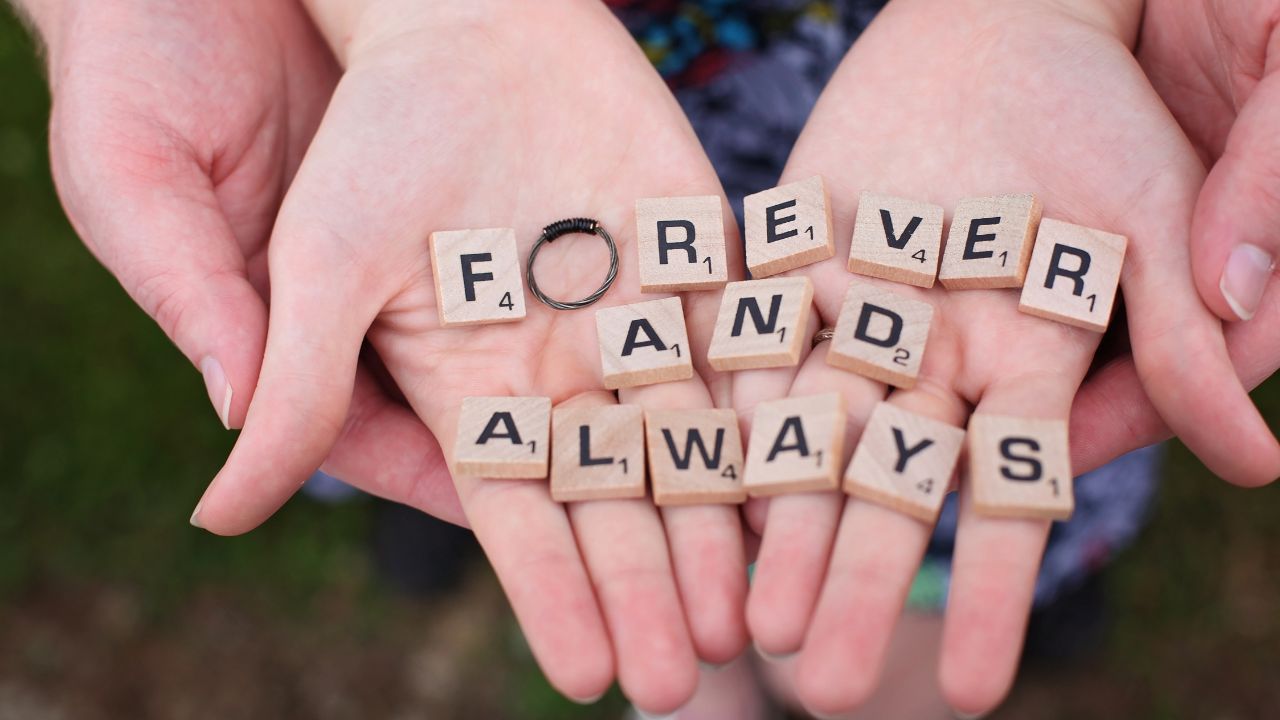 Two hands holding Scrabble tiles that spell "forever and always," with an engagement ring on one of the tiles, symbolizing romantic commitment and happy engagement.