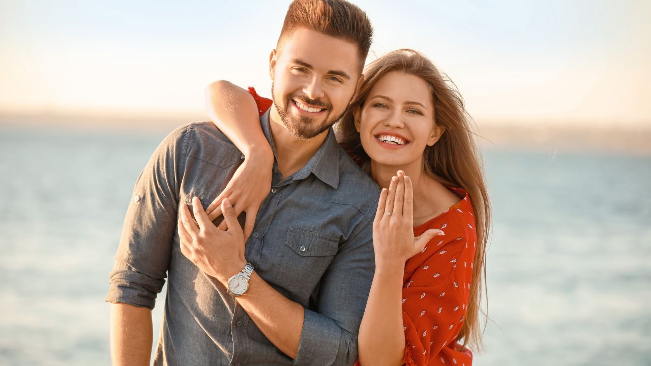 A joyful couple embracing by the sea at sunset; the man in a blue shirt and the woman in a red blouse, both smiling warmly at the camera, celebrating their happy engagement.