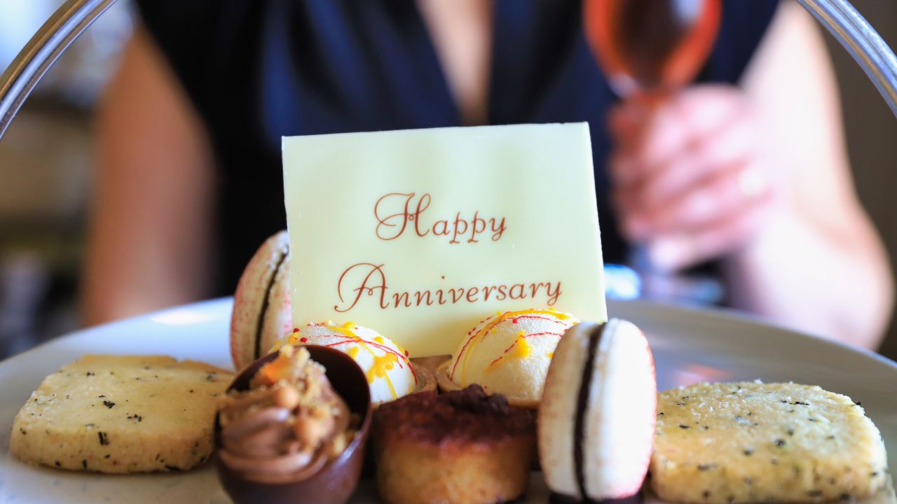 A close-up of a plate bearing a "wedding anniversary wishes for wife" card, surrounded by an assortment of desserts including macarons and cookies, with a person holding a wine glass in the