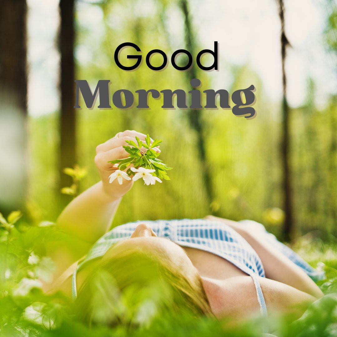 A peaceful good morning image featuring a woman lying on her back in a lush green meadow, holding a small bouquet of white flowers. The text "Good Morning" is displayed in a charming font at the top. The background is filled with vibrant greenery and trees, creating a serene and relaxing nature scene perfect for a gentle morning greeting.