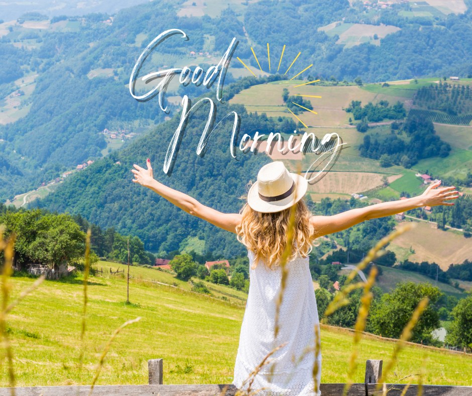 A vibrant good morning image featuring a woman in a white dress and sun hat, standing with her arms outstretched, overlooking a picturesque valley with lush green hills and fields. The text "Good Morning" is elegantly written in white with a sun illustration. The image captures the beauty and tranquility of the countryside, perfect for a refreshing morning greeting.