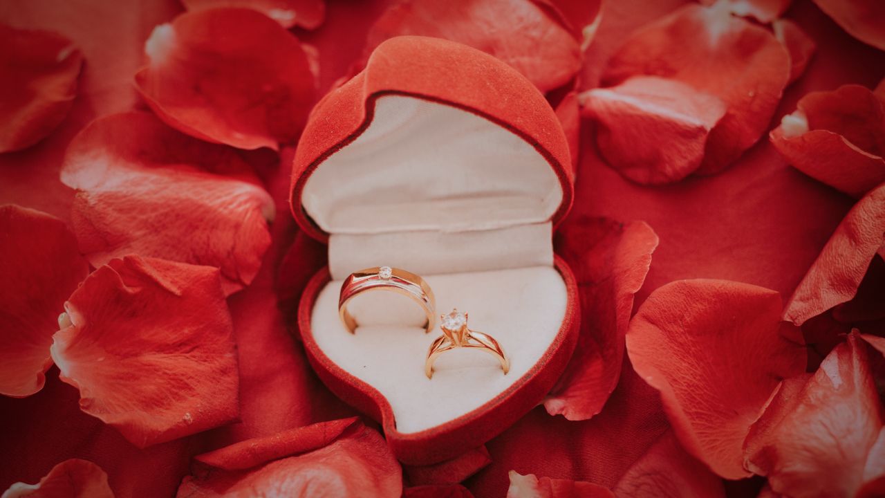 Two engagement rings in a heart-shaped box surrounded by red rose petals, evoke happy anniversary wishes for a husband. One ring features a solitaire diamond; the other is simpler with a small stone, set