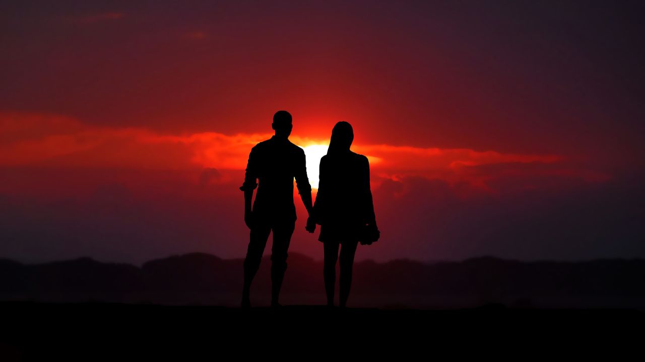 Silhouette of a couple holding hands against a vibrant red sunset with a dramatic cloud-filled sky, capturing the essence of Happy Monthsary Messages.