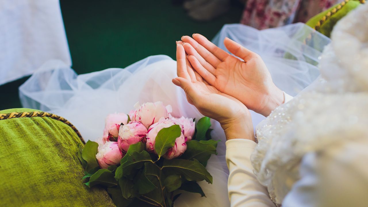 Close-up of a bride's hands cupped together next to a bouquet of pink peonies, showing elegant white sleeves and details of her dress, with a hint of green ambient background, capturing the