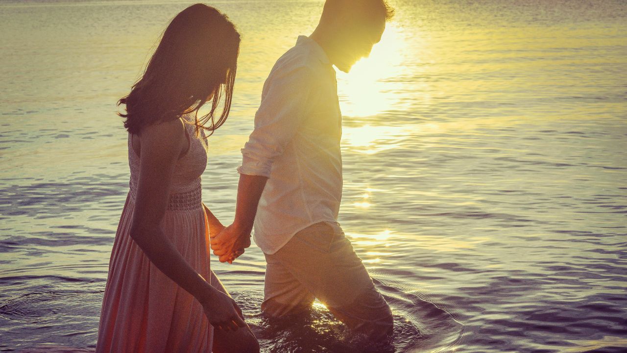 A silhouette of a couple holding hands while walking in shallow water at sunset, with the sun low on the horizon casting a warm glow over the scene, perfect for celebrating a happy monthsary.