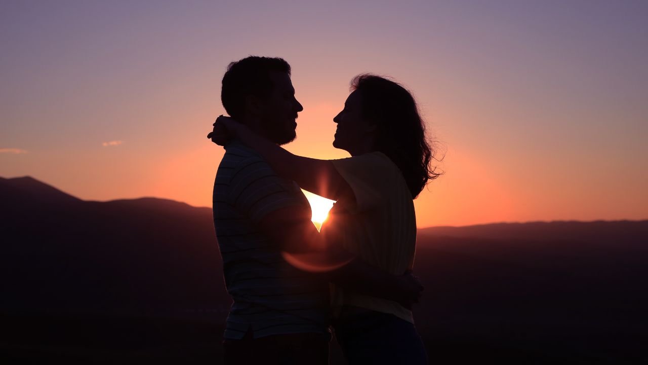 A silhouette of a boyfriend and girlfriend embracing against a sunset backdrop with vibrant orange and purple hues casting a warm glow over a mountainous horizon.