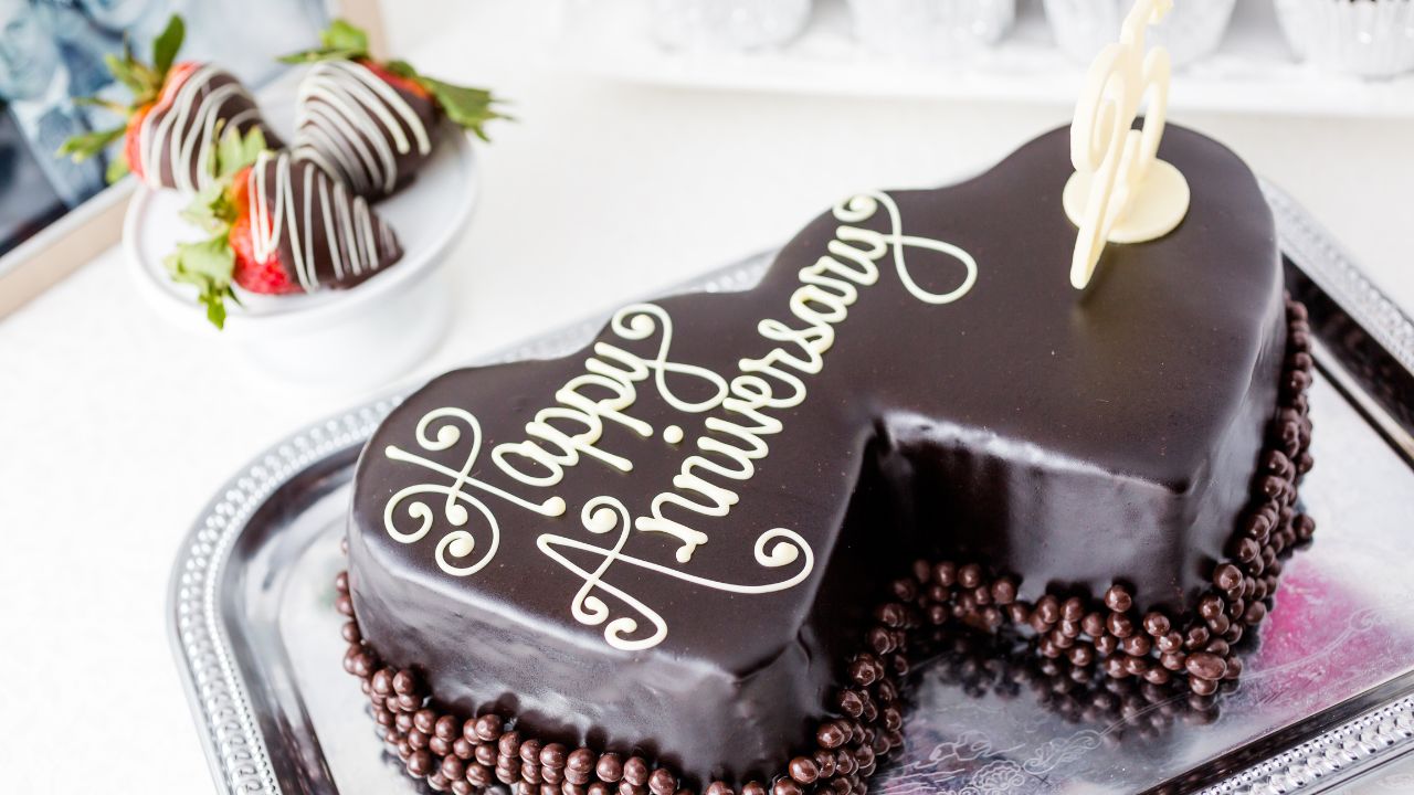 A heart-shaped chocolate cake with "wedding anniversary wishes for wife" written in cursive icing, garnished with chocolate pearls and chocolate-dipped strawberries on a silver tray.
