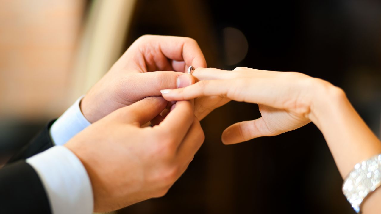 A groom placing an engagement ring on a bride's finger, both dressed in formal attire, with a focus on their hands against a blurred background.