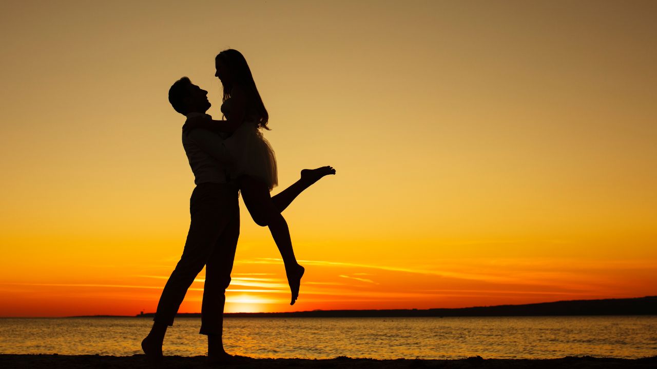 Silhouette of a boyfriend and girlfriend embracing and lifting up in joy against a sunset over the ocean, with warm orange tones illuminating the horizon.