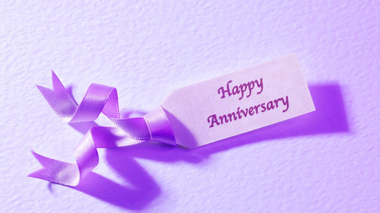 A purple ribbon and a tag with "wedding anniversary wishes for wife" written on it, placed on a textured purple background.