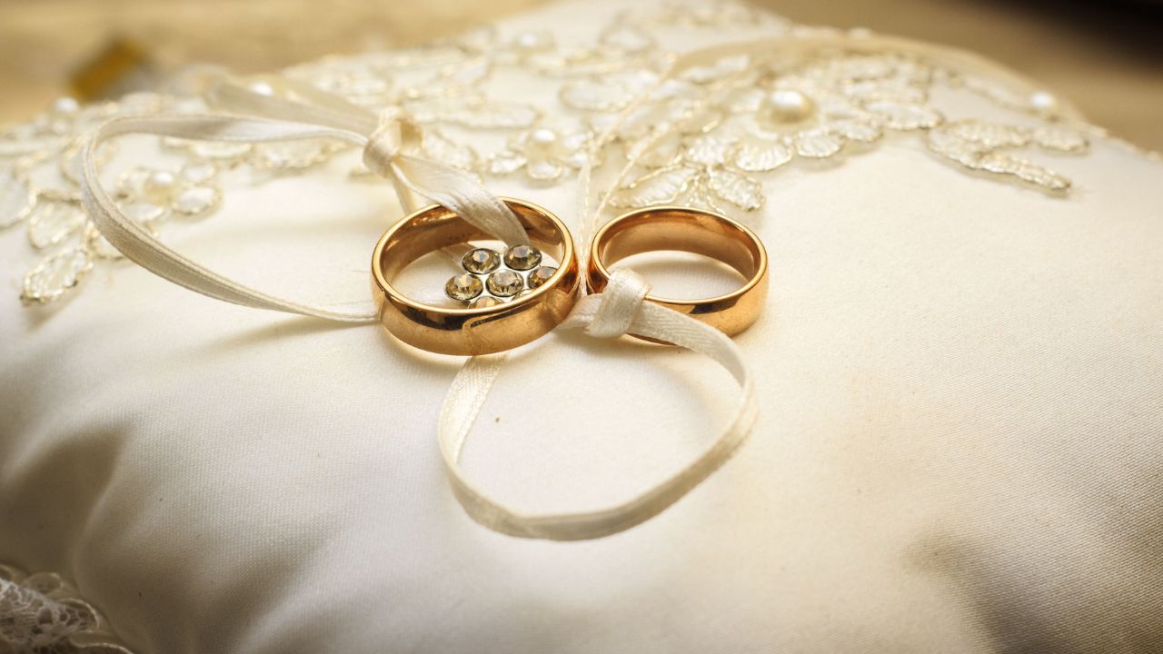 Two golden engagement rings tied with a thin ribbon, resting on a white lace pillow embellished with pearls and beads.