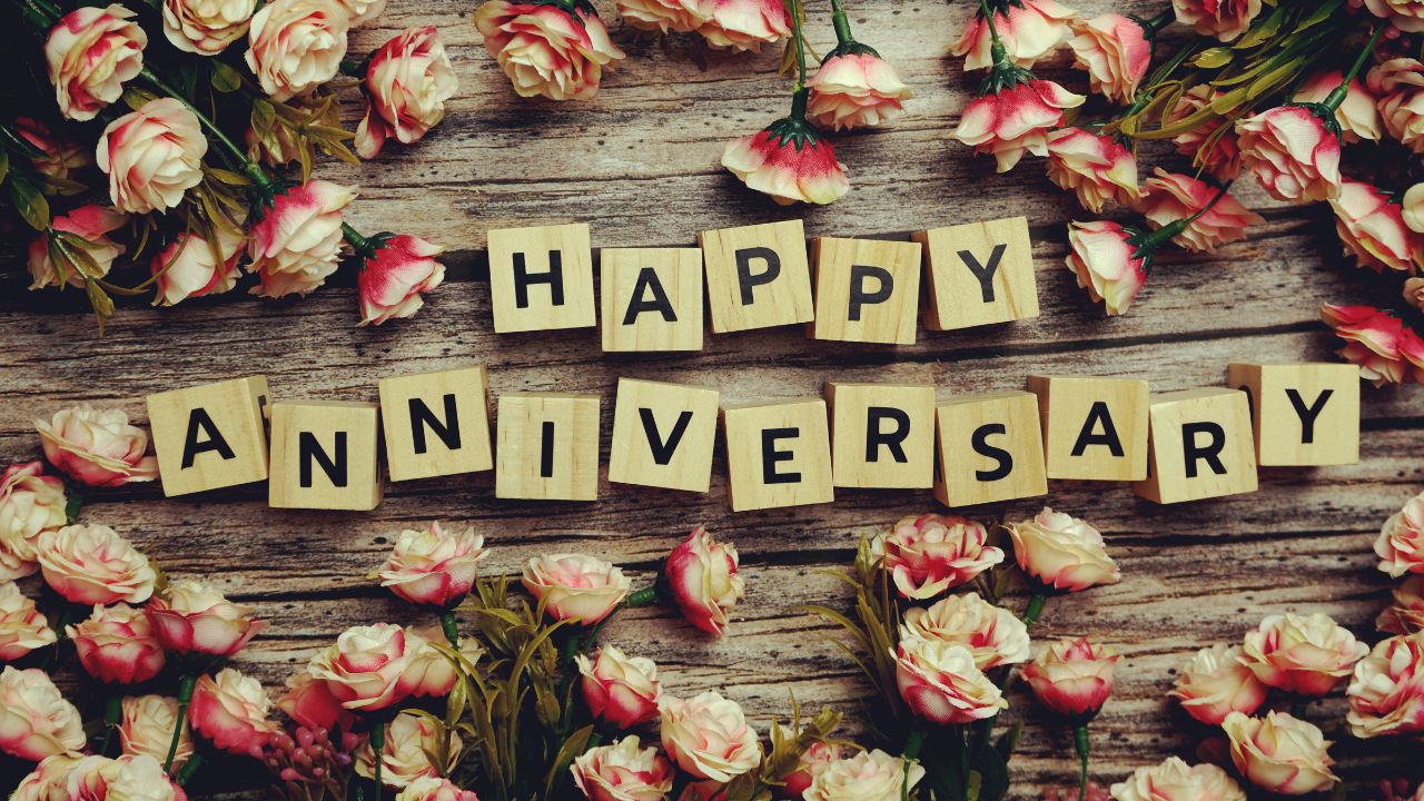 The words "wedding anniversary wishes for wife" spelled out in wooden block letters surrounded by scattered pink and white roses on a rustic wooden background.