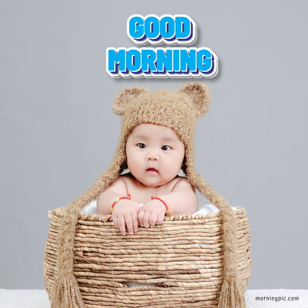 A baby wearing a cozy bear hat peers over the rim of a woven basket against a gray background. Above the baby, the text reads "GOOD MORNING" in bright, cheerful blue letters.